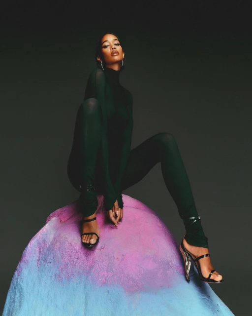 Adult female in stylish green bodysuit and high-heel sandals sits on a colorful stone-like form against a dark gray studio backdrop.