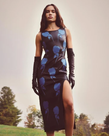 Model in dark sleeveless dress with blue floral motifs and black leather gloves standing outdoors on grassy hill
