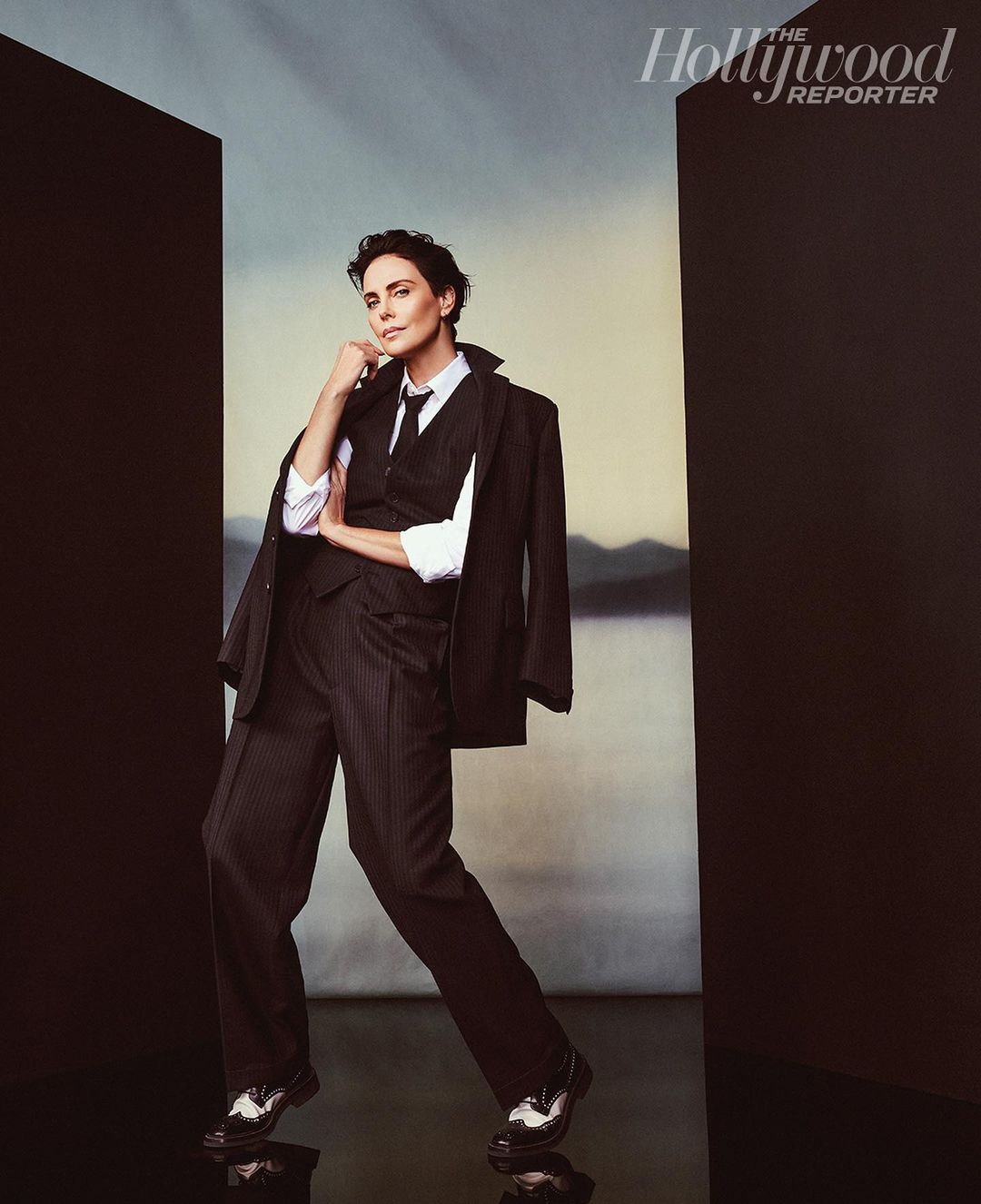 Charlize Theron poses in a dark pinstripe suit, vest, and tie in a Hollywood Reporter studio portrait. Her hand rests on her chin.