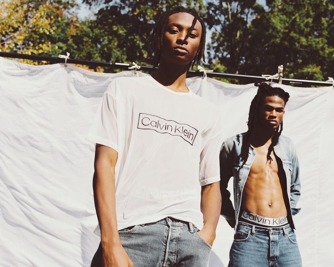 Two adult males pose outdoors in Calvin Klein apparel against a draped white backdrop and green trees, showcasing casual fashion.