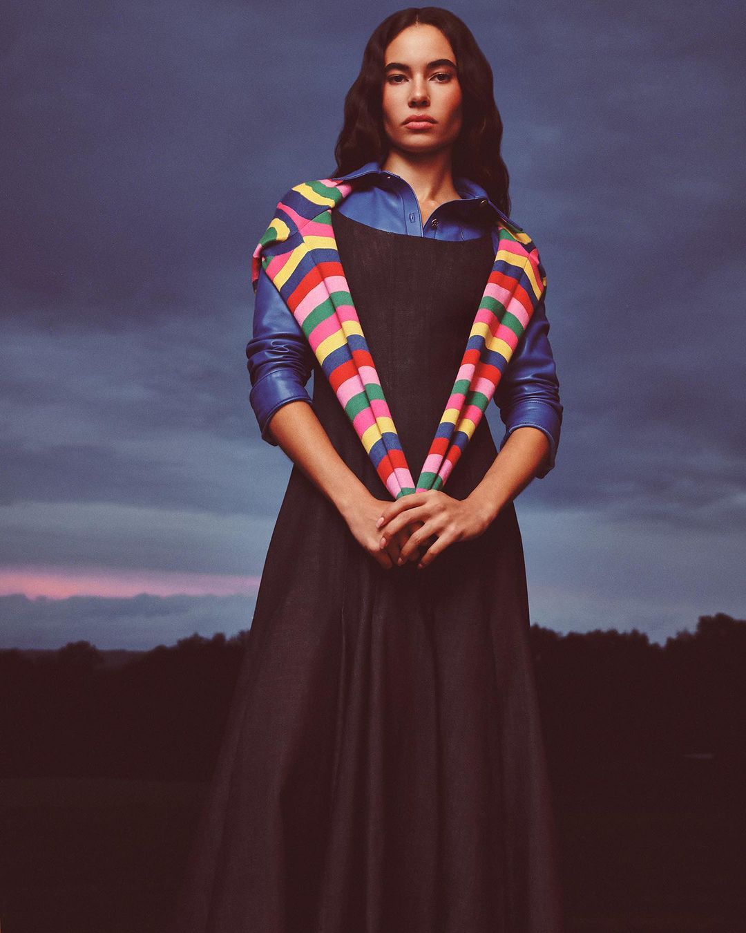 Woman in dark long dress with blue shirt and striped colorful scarf outdoors against a twilight sky