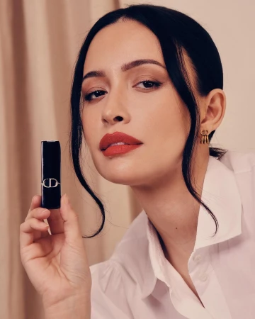 Woman in white shirt holding a black lipstick tube while wearing bold red lipstick and gold earrings against a neutral backdrop.