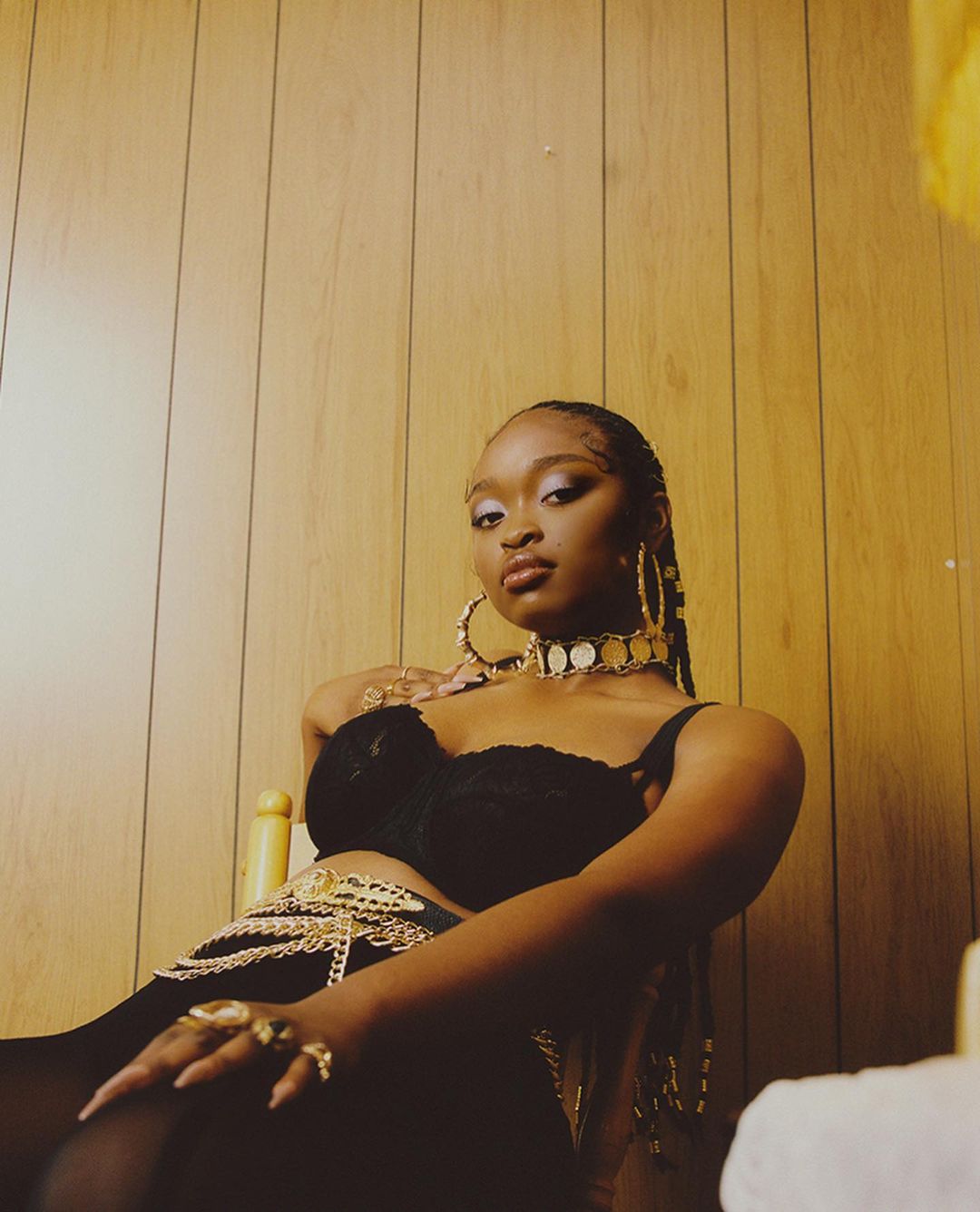 Portrait of a woman wearing black top and gold jewelry against wood panel wall. She's indoors with neutral and yellow lighting.