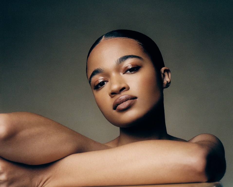 Portrait of a young adult woman with dark hair and skin, light brown lipstick, and arms folded, set against a soft neutral backdrop.