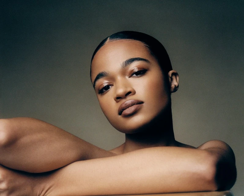 Portrait of a young adult woman with dark hair and skin, light brown lipstick, and arms folded, set against a soft neutral backdrop.