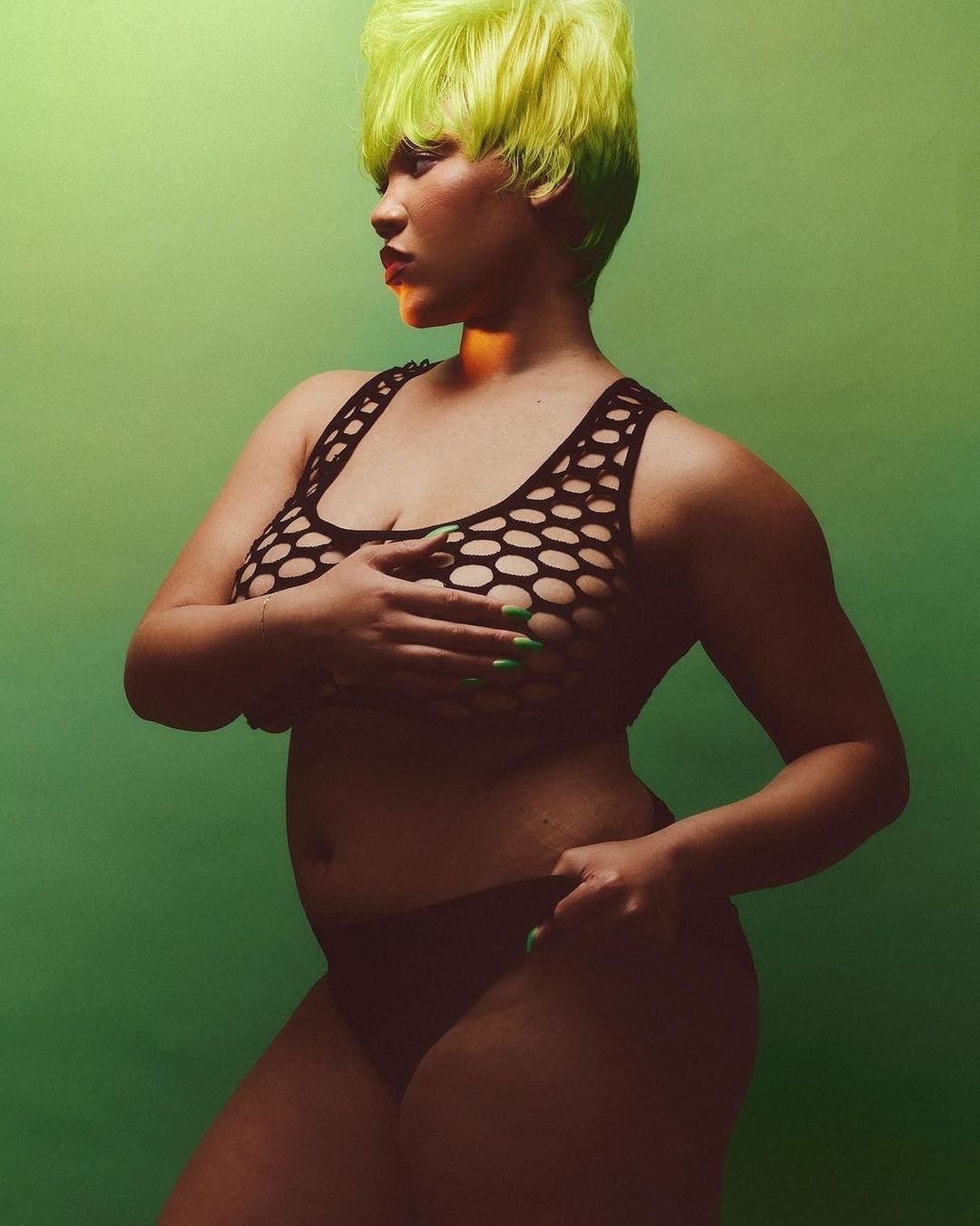 Portrait of a plus-size woman with neon green hair and black lingerie posing against a green studio backdrop with soft, diffused lighting.