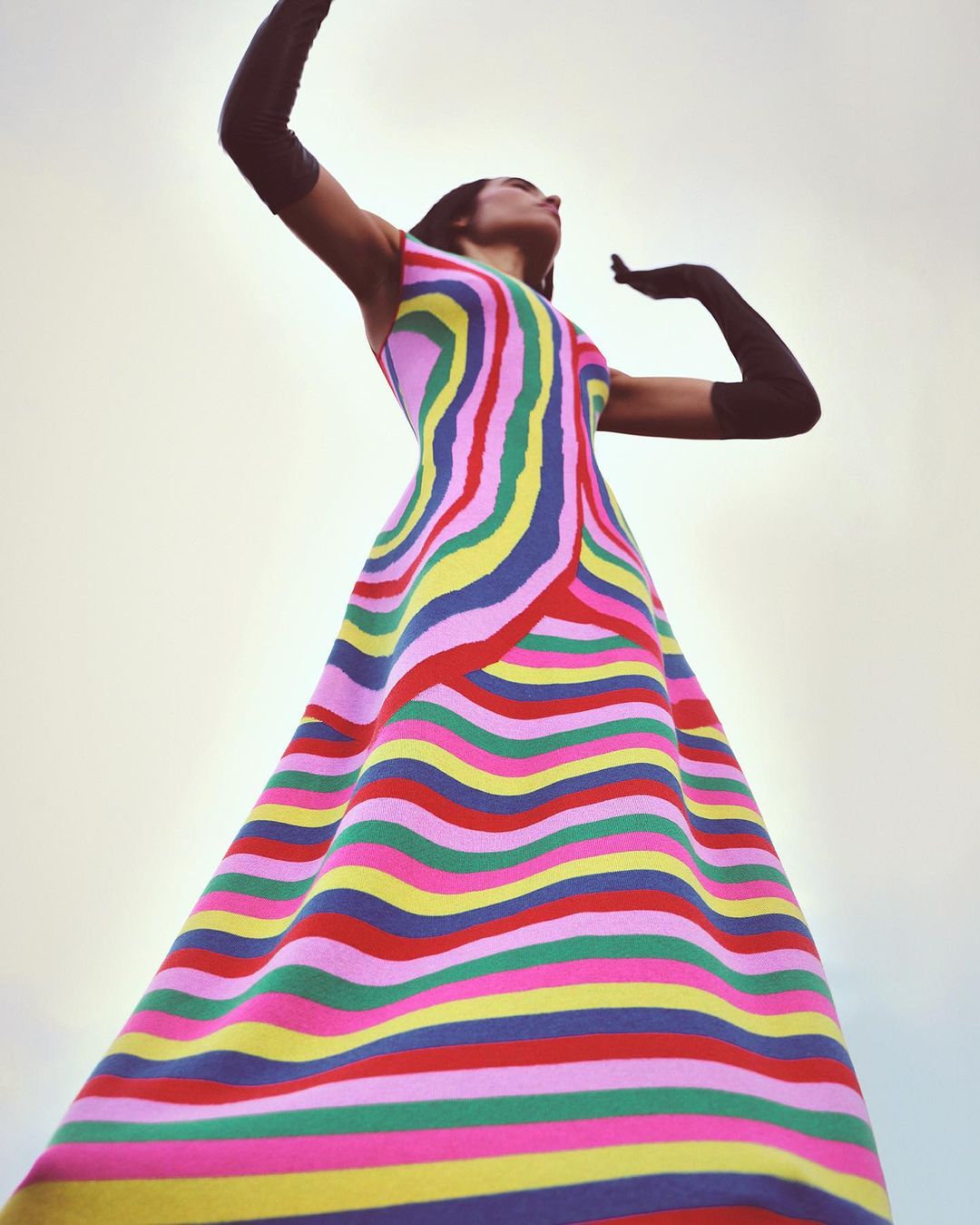 Woman in colorful striped dress and long gloves poses against a bright sky; low angle shot emphasizing the dress and pose.