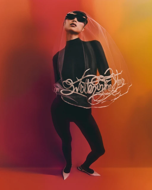Fashionable person in black sunglasses, and a black suit with a bridal veil that has text on it, posing against a multicolored backdrop.