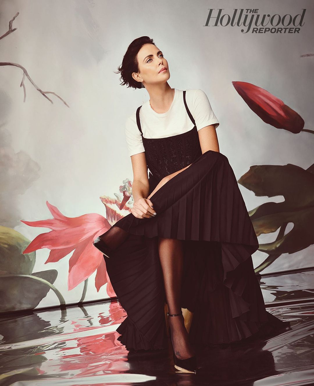Charlize Theron poses seated in a dark dress with a white t-shirt, black pumps, and sheer stockings, with muted, floral, water-like background.
