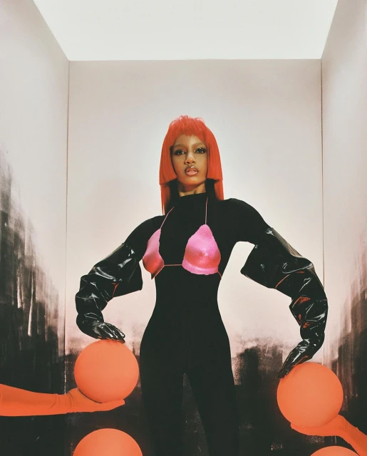 Woman with red hair poses in a black bodysuit and pink bra with orange spheres in a white and black graphic studio backdrop.