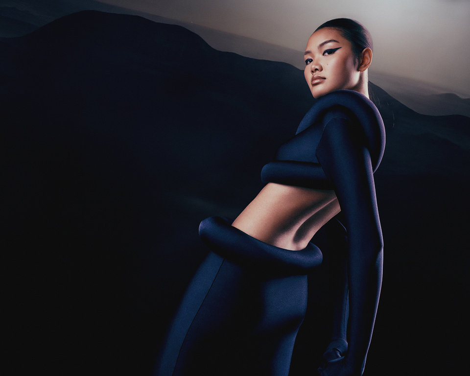 Fashionable adult woman in a dark blue bodysuit poses confidently against a blurred mountain backdrop, showcasing modern clothing and style.