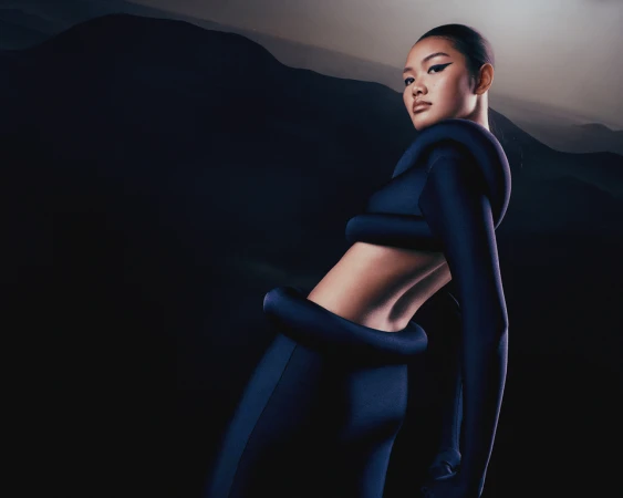 Fashionable adult woman in a dark blue bodysuit poses confidently against a blurred mountain backdrop, showcasing modern clothing and style.