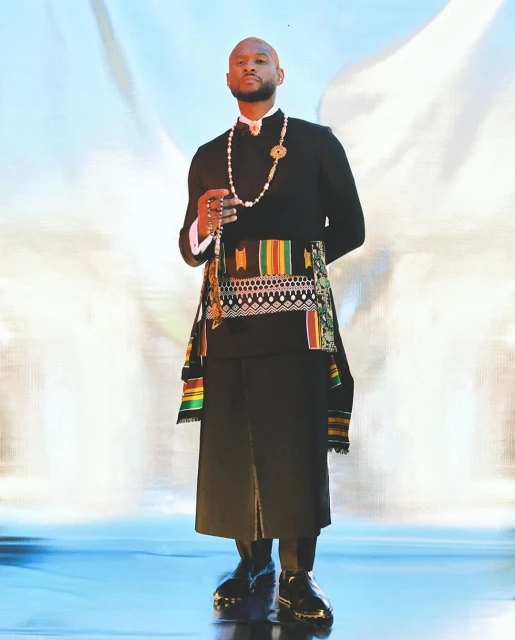 Bald adult male fashion model poses in black suit with colorful sash and pearls in a full shot against a blurred gradient backdrop.
