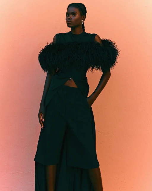 Elegant dark-skinned woman in a black, feathered, cold-shoulder dress poses against a peach backdrop. She is looking slightly to the left.
