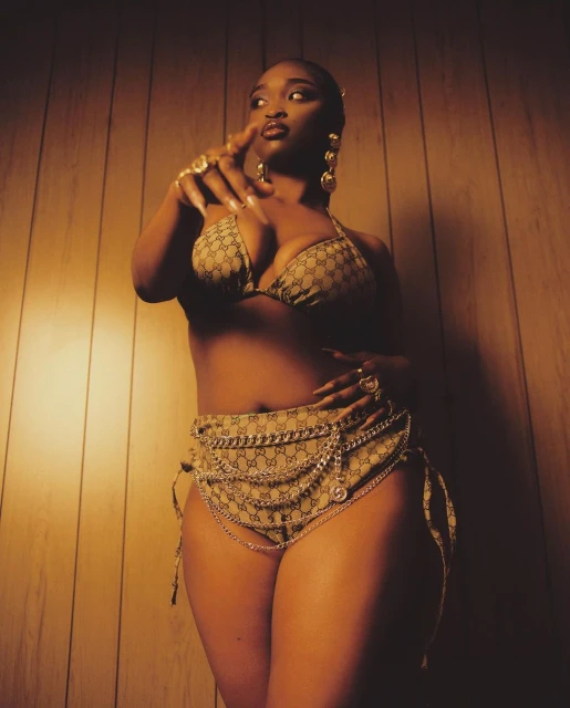 A dark-skinned woman poses in a patterned bikini and skirt with gold jewelry against a warm-toned wall, pointing her finger at the camera.