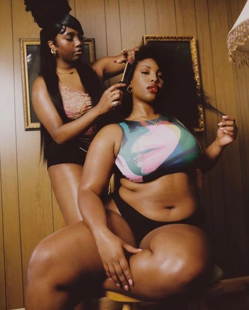 Two African-American women indoors. One woman sitting on stool while another stands behind her combing her hair in a room with wood panelling.