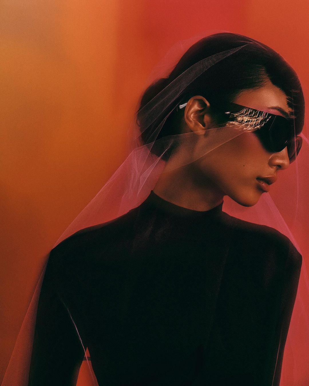 Woman in a black turtleneck and avant-garde sunglasses poses against a gradient background. Tulle drapes over her head and shoulders.