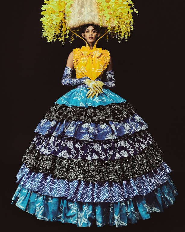 Woman poses in a tiered, printed gown with a floral headdress against a dark backdrop, showcasing fashion and elaborate design.