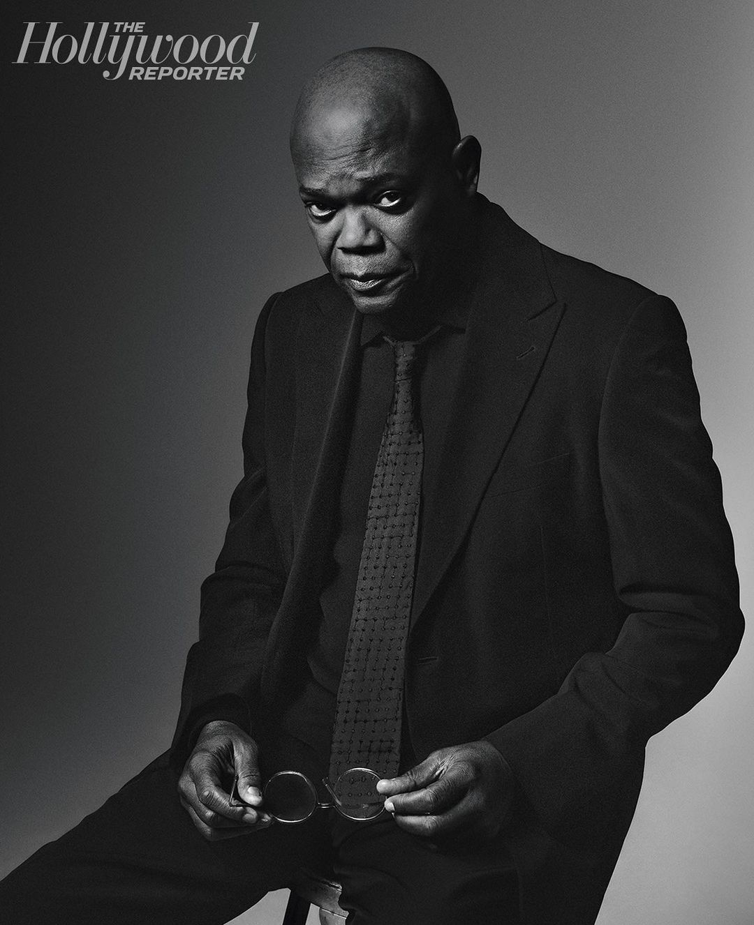 Portrait of Samuel L. Jackson in a dark suit and tie, seated and holding eyeglasses against a grey gradient backdrop, with The Hollywood Reporter logo.