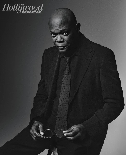Portrait of Samuel L. Jackson in a dark suit and tie, seated and holding eyeglasses against a grey gradient backdrop, with The Hollywood Reporter logo.