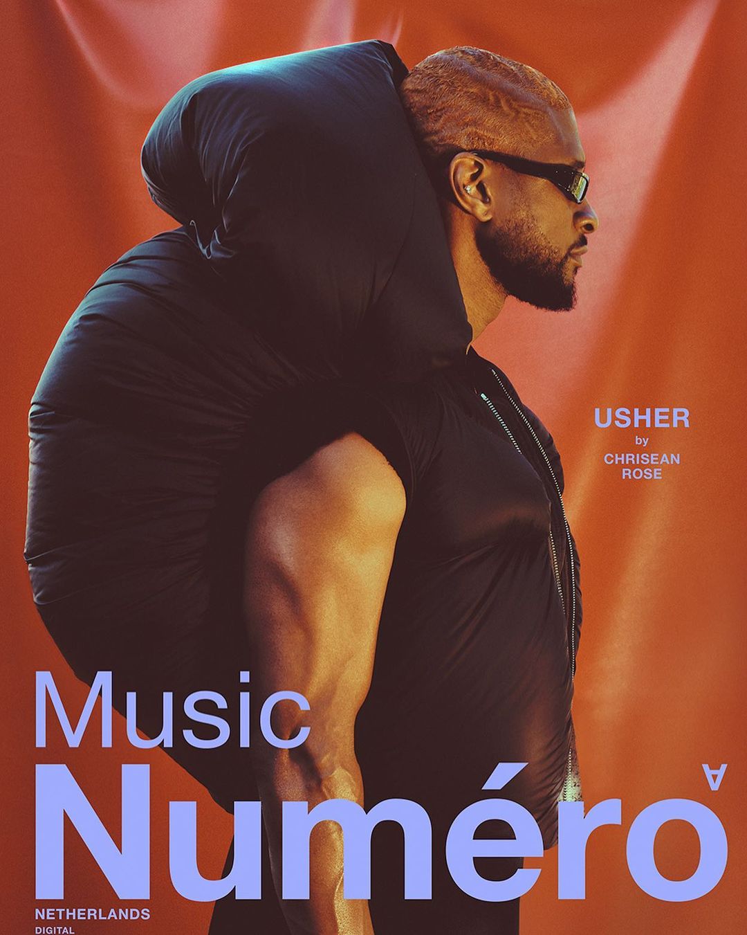 Singer Usher is on a magazine cover in orange tones, wearing black glasses and a sleeveless top, and carrying a puffy black wrap.
