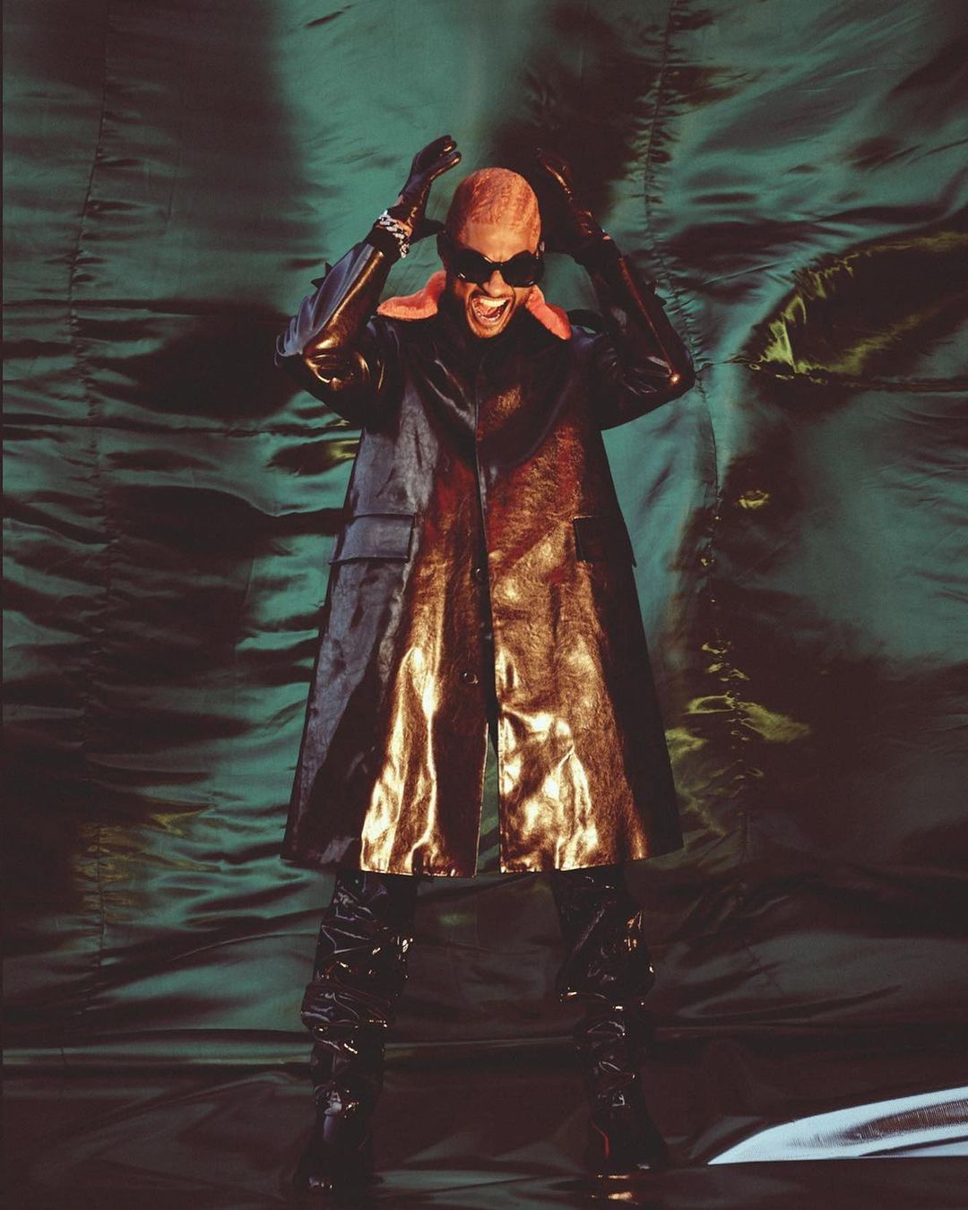 Adult standing with hands up, wearing sunglasses, patent leather boots and coat, in front of a textured green backdrop.