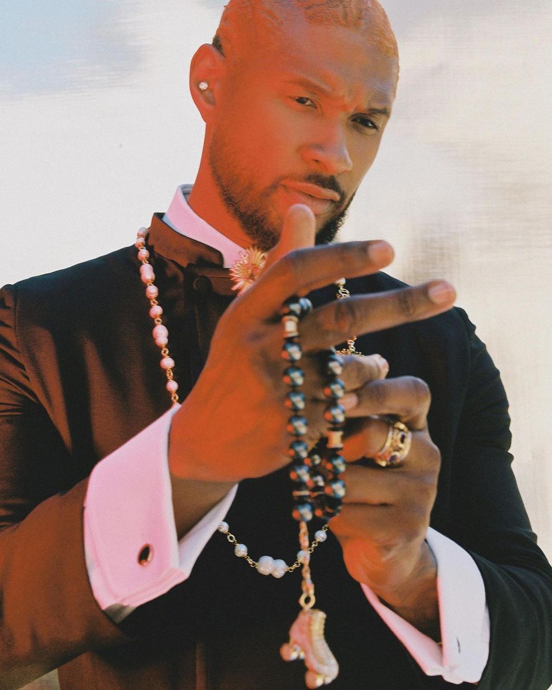 Portrait of an adult male with pale hair wearing a black tuxedo, white shirt, and pearls. He is holding a black beaded necklace.