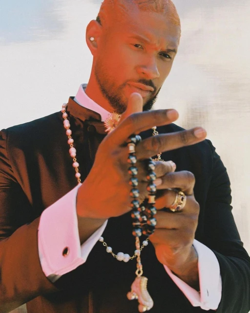 Portrait of an adult male with pale hair wearing a black tuxedo, white shirt, and pearls. He is holding a black beaded necklace.
