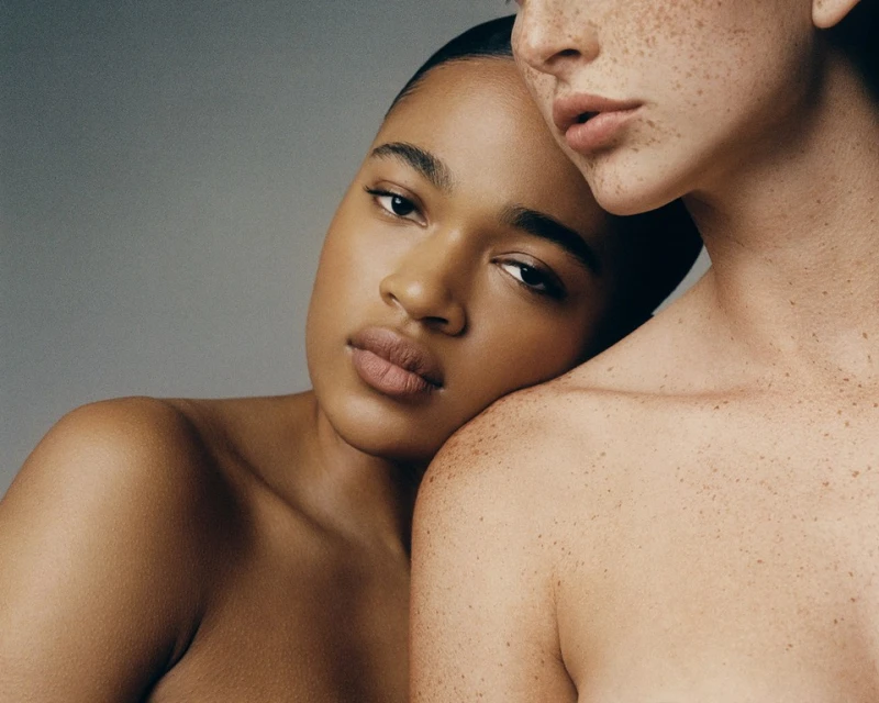 Close-up studio portrait of two women with clear skin in front of a solid gray background. One woman rests her head on the other's shoulder.
