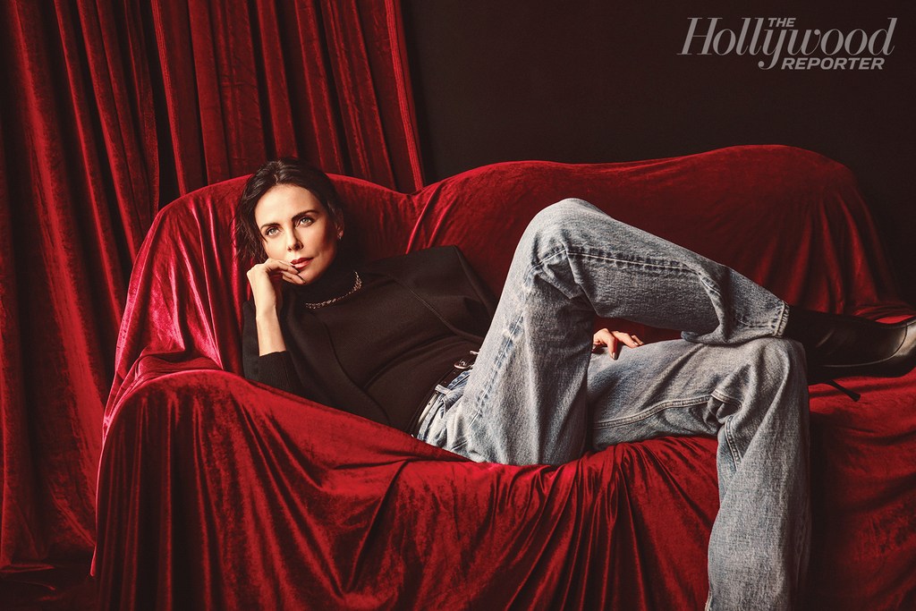 Charlize Theron lounges on a red velvet sofa, posing for a Hollywood Reporter portrait in jeans, a black turtleneck, and a black blazer.