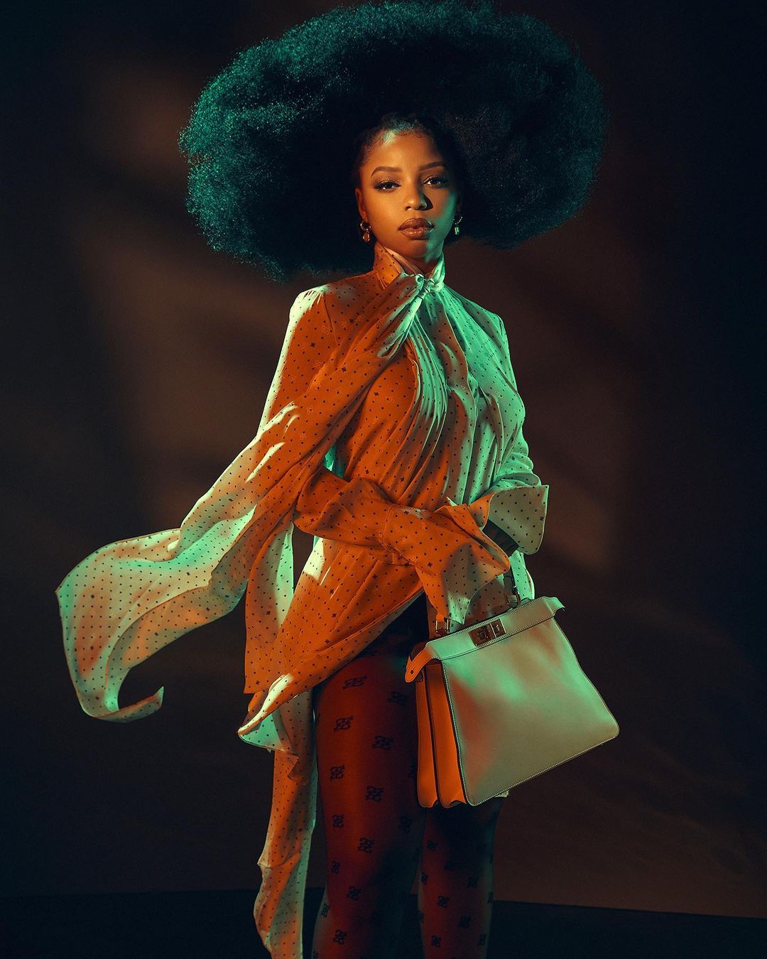 Woman with large afro poses in polka dot top and patterned leggings with a white satchel purse in a studio setting.