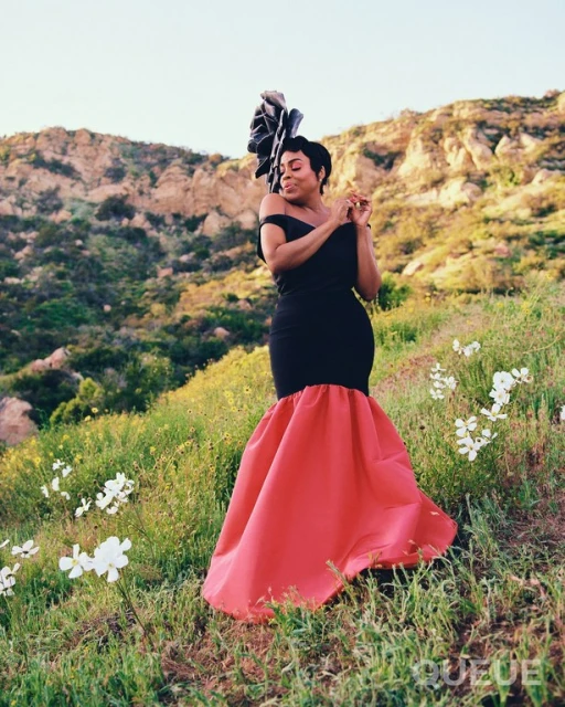 A woman in a black and red dress stands in a field with hills in the background, wearing a large floral headpiece and gesturing with her hands.