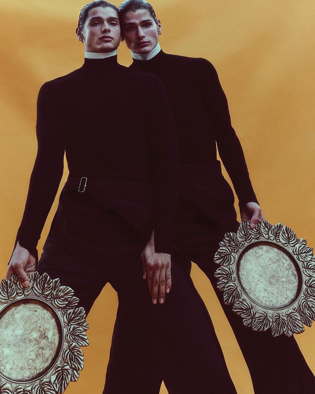 Two men in black turtlenecks pose against a yellow background, each holding a decorative silver plate, creating a symmetrical fashion image.