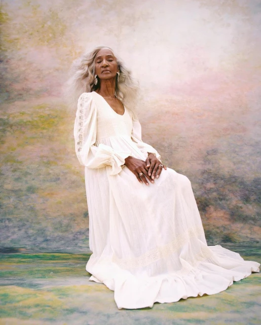 Elegant elderly Black woman with long gray hair sits in a vintage white dress against a soft watercolor backdrop in a portrait shot.