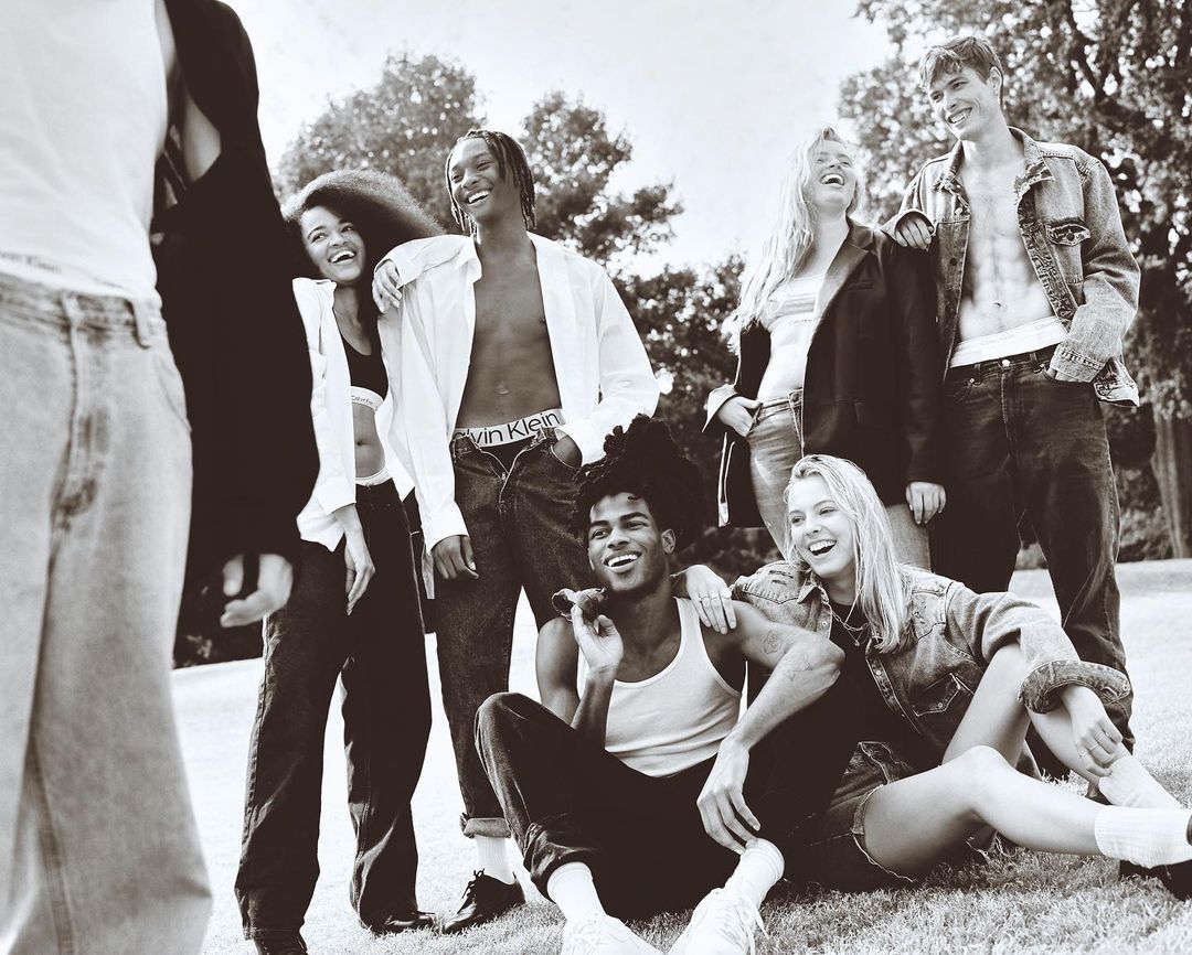 Group of young adult friends in casual wear, jeans, and light tops, laughing and relaxing together on a grassy hill against a sunny sky.