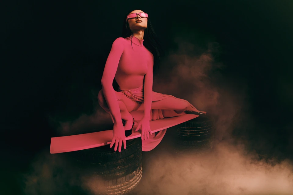 A woman in pink bodysuit and glasses crouches on a platform atop tires amid smoke, with a dark backdrop, fashion-forward and stylized.
