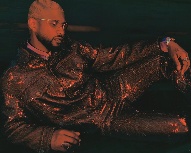 A fashionable adult male with platinum blonde hair and glasses poses in a sequined denim suit indoors under moody lighting.