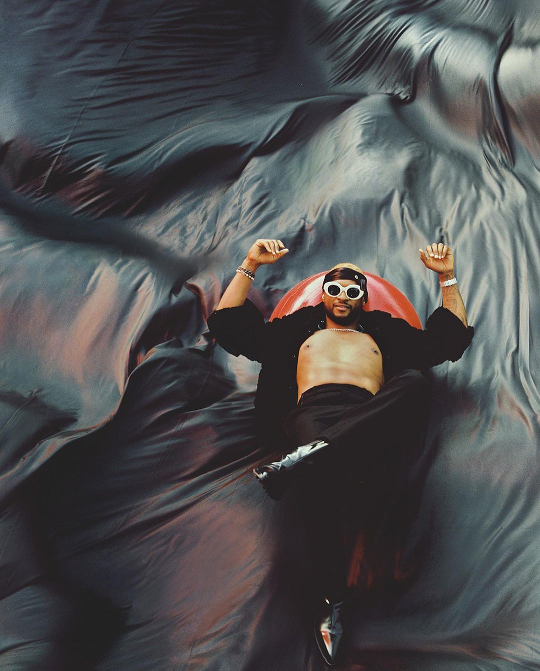 Man lying on a reflective surface wearing sunglasses, open shirt, and black pants with shiny shoes against a flowing fabric backdrop.