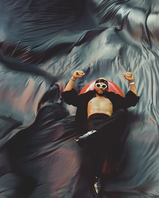 Man lying on a reflective surface wearing sunglasses, open shirt, and black pants with shiny shoes against a flowing fabric backdrop.
