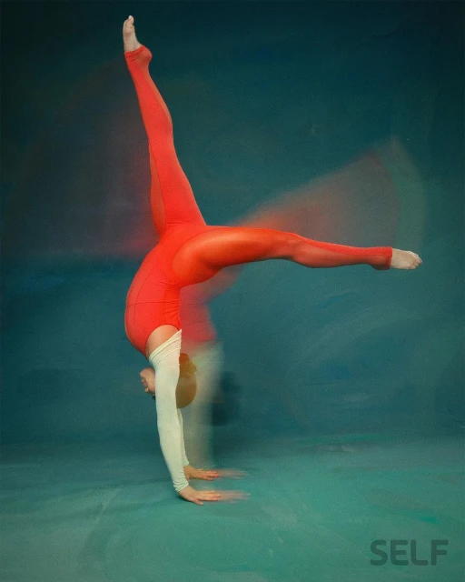 A woman in a red bodysuit is doing a handstand with her legs split in the air against a teal background in a fitness pose.