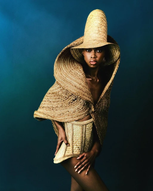 A stylish dark-skinned woman poses in straw-woven couture hat and corset against a teal studio backdrop, creating a striking editorial image.