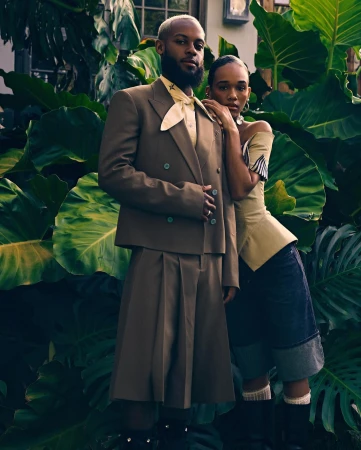 Portrait of a black man in a brown suit and a black woman posing outdoors surrounded by lush green tropical foliage.