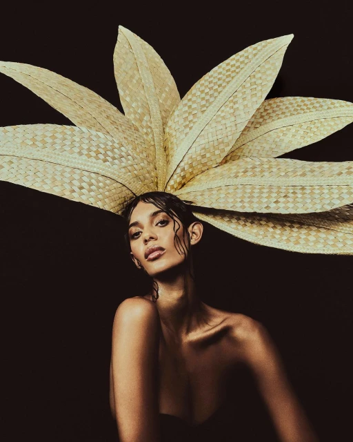 Portrait of a woman with dark hair wearing a tan woven headpiece that resembles leaves against a dark background, beauty-style lighting.