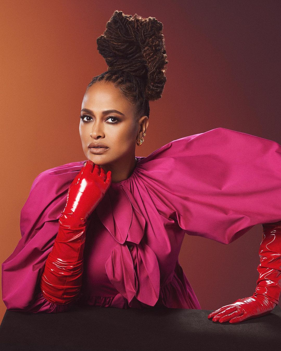 Glamorous studio portrait of a woman with an elaborate hairstyle and bright pink dress wearing long, shiny red gloves.