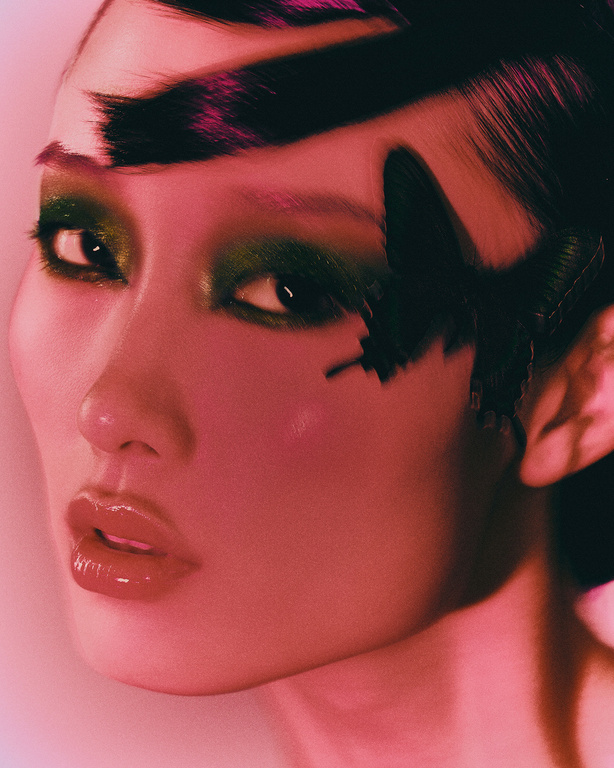 Glamorous portrait of an Asian woman with green eyeshadow, glossy lips, and a butterfly embellishment in a pink studio environment.
