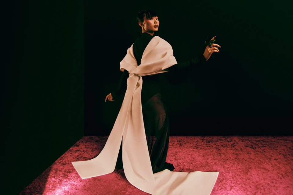 Woman in dark dress with large ribbon, posing in a photo studio. The model is highlighted against a dark backdrop with a pink floor.