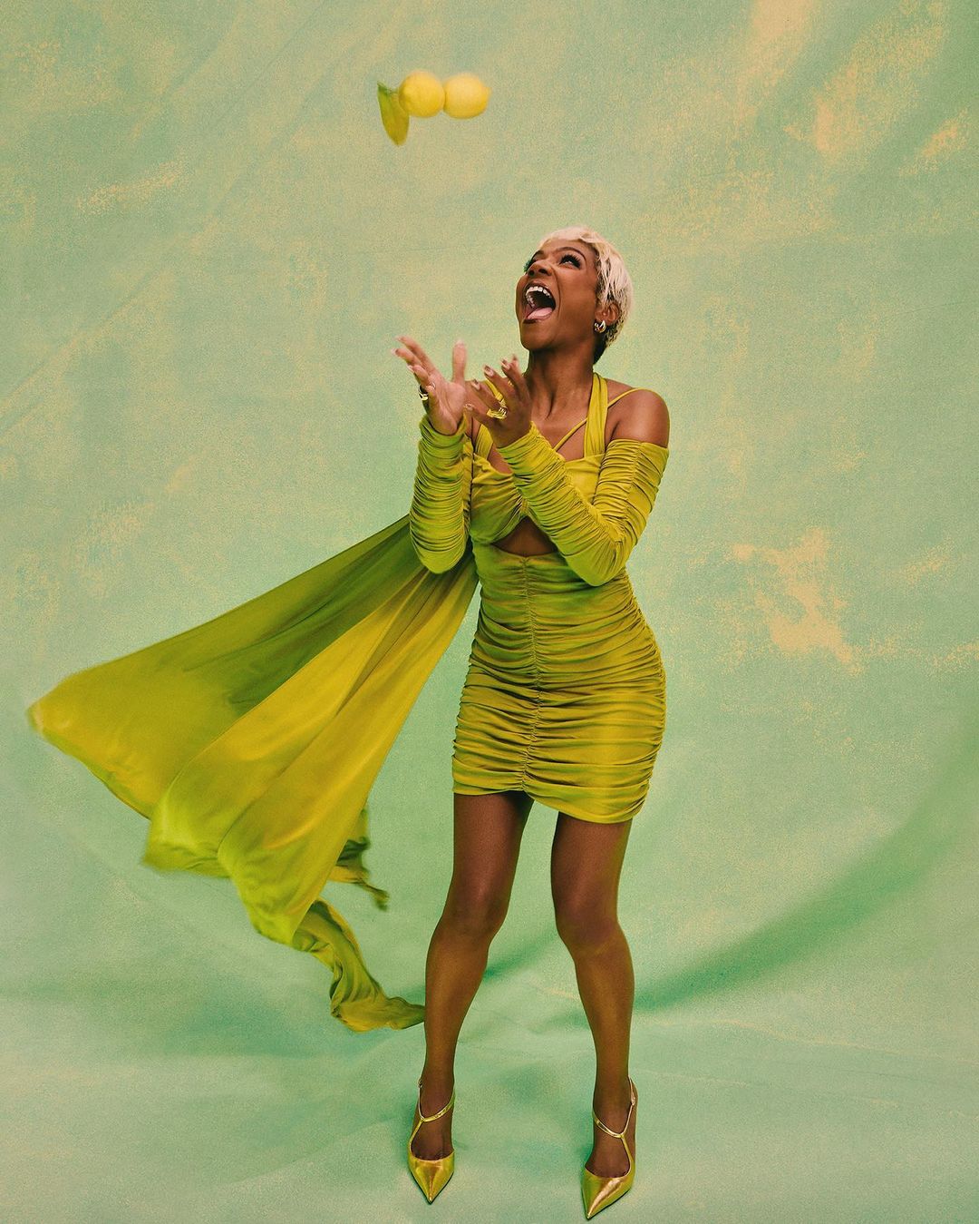 An adult woman in a yellow dress laughs with open arms as she looks up at lemons above her in a light green indoor studio setting.