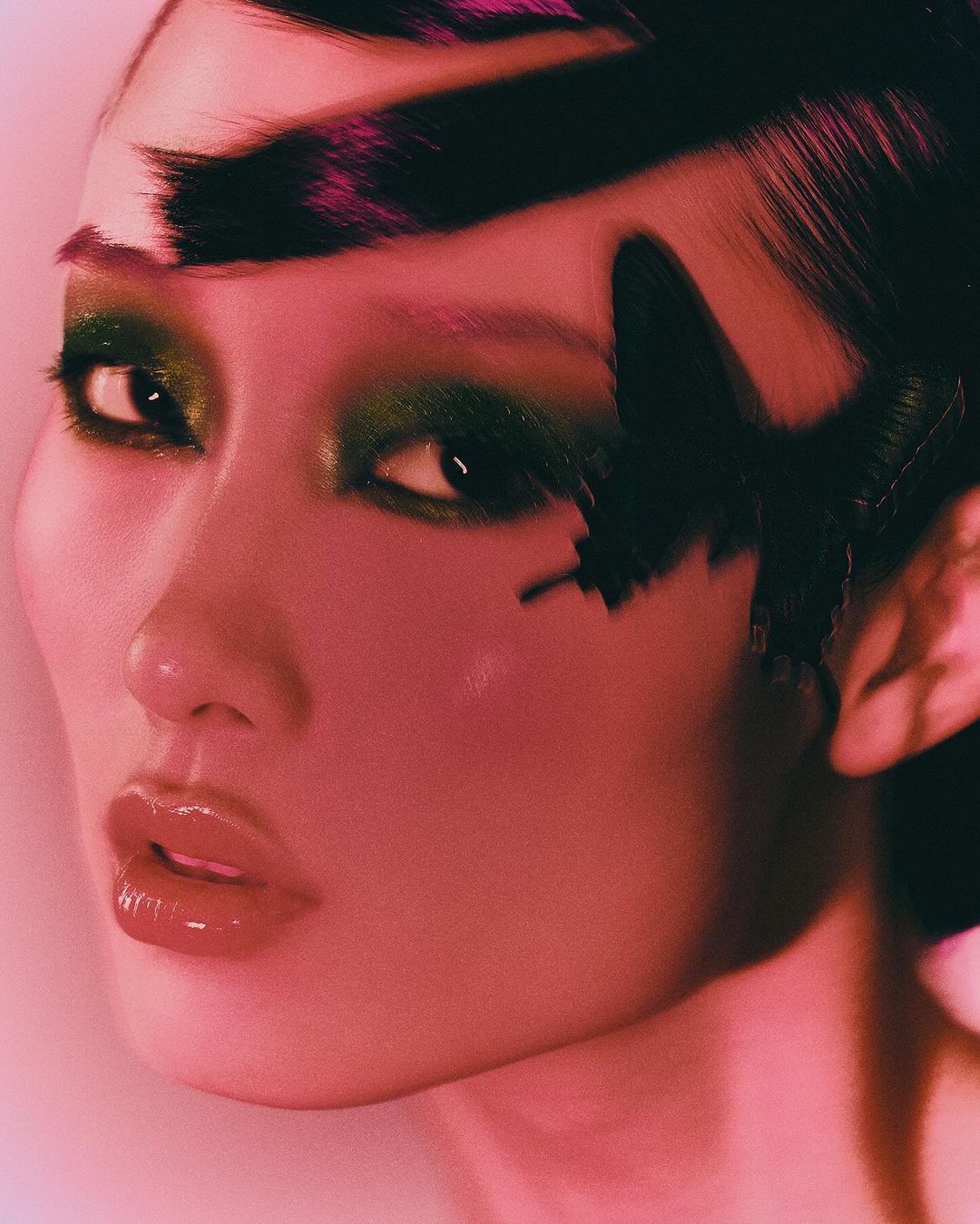 Close-up studio shot of a woman with dark hair, heavy green eye makeup, and a black butterfly accessory near her eye. Pink-toned lighting.