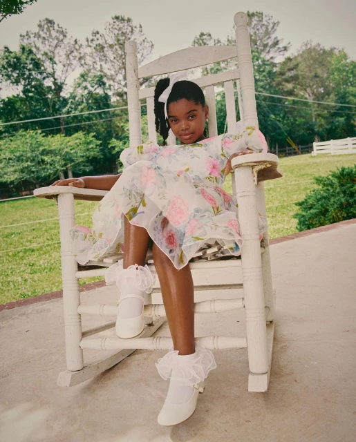 A young African-American girl in a white floral dress and white shoes poses sitting in a white rocking chair outside on a sunny day.