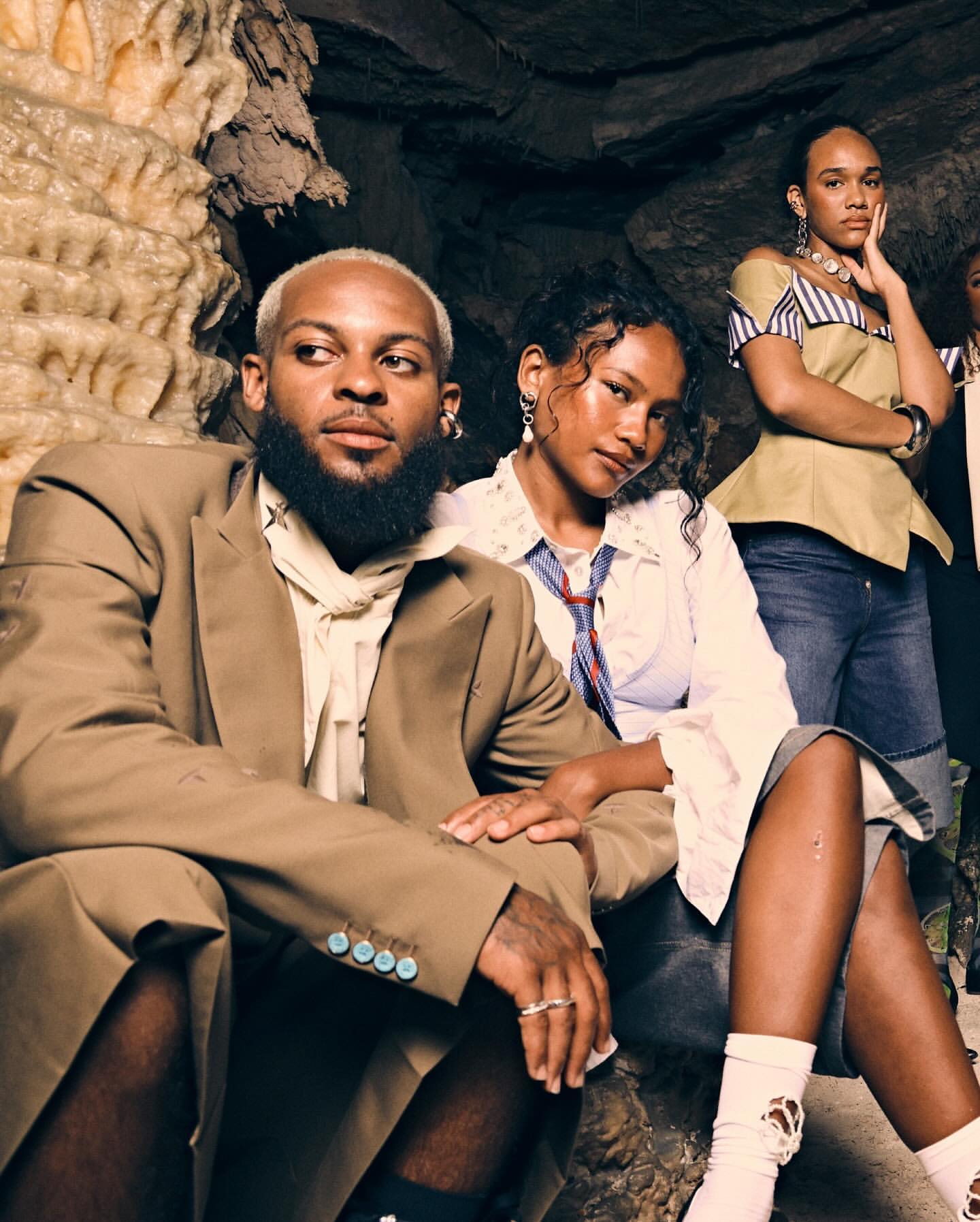 Group of four people sitting in a cave wearing stylish clothing and jewelry, posing for a portrait-style fashion shot.